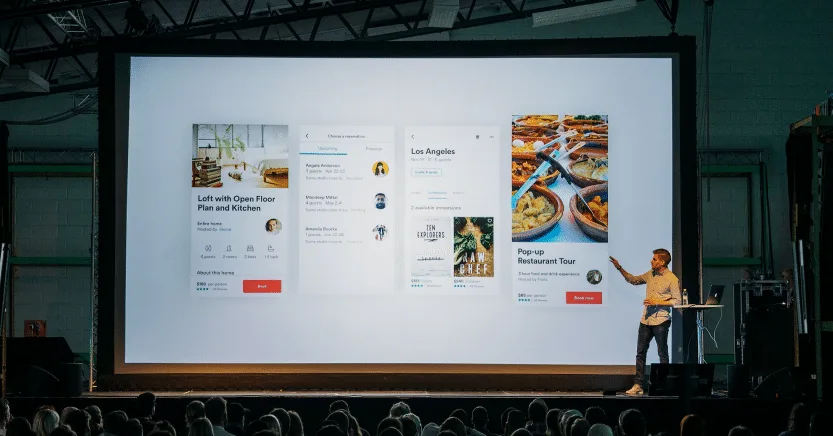 Man presenting mobile app mockups on large screen to audience in dark auditorium.